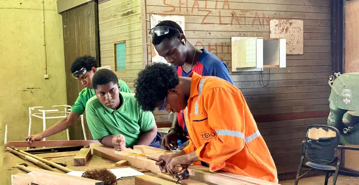 A group of individuals working on a woodworking project in a workshop. The workspace includes a large wooden table with tools, wooden planks, and other materials scattered across it. One person in an orange safety uniform is actively measuring or marking wood, while others observe and assist. The background features workshop equipment, such as a machine and chairs, along with a wooden wall bearing markings and notes.