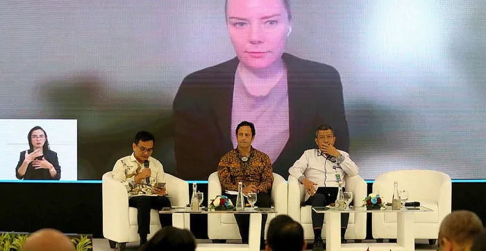 Three persons sitting on stage with a screen on the back featuring another speaker