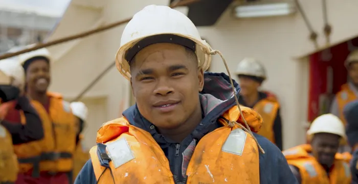A young seafarer wearing a safety helmet and orange life vest smiles at the camera aboard a fishing vessel in South Africa. Behind him, fellow crew members in similar safety gear prepare for work. The photo was taken as part of the ILO’s 8.7 Accelerator Lab Initiative, which supports decent work and improved labour conditions in the fishing sector.